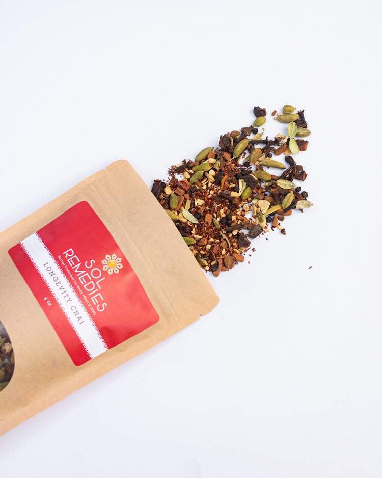 Brown kraft paper pouch of ‘Sol Remedies Longevity Chai’ tea lying on its side, with colorful loose tea leaves and spices spilling out onto a white background