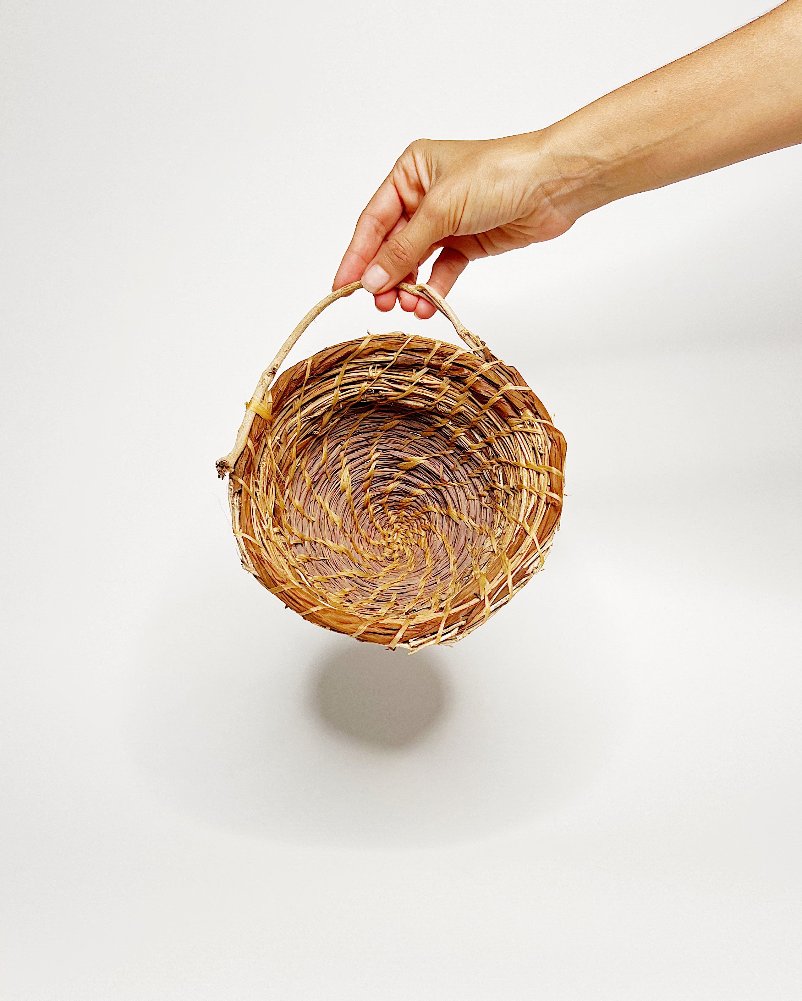 Handwoven basket crafted from lemongrass, lauaʻe fern, morning glory vine, and hau fiber, featuring a natural spiral weave pattern and a small organic handle, displayed against a clean white background. Hand holding basket