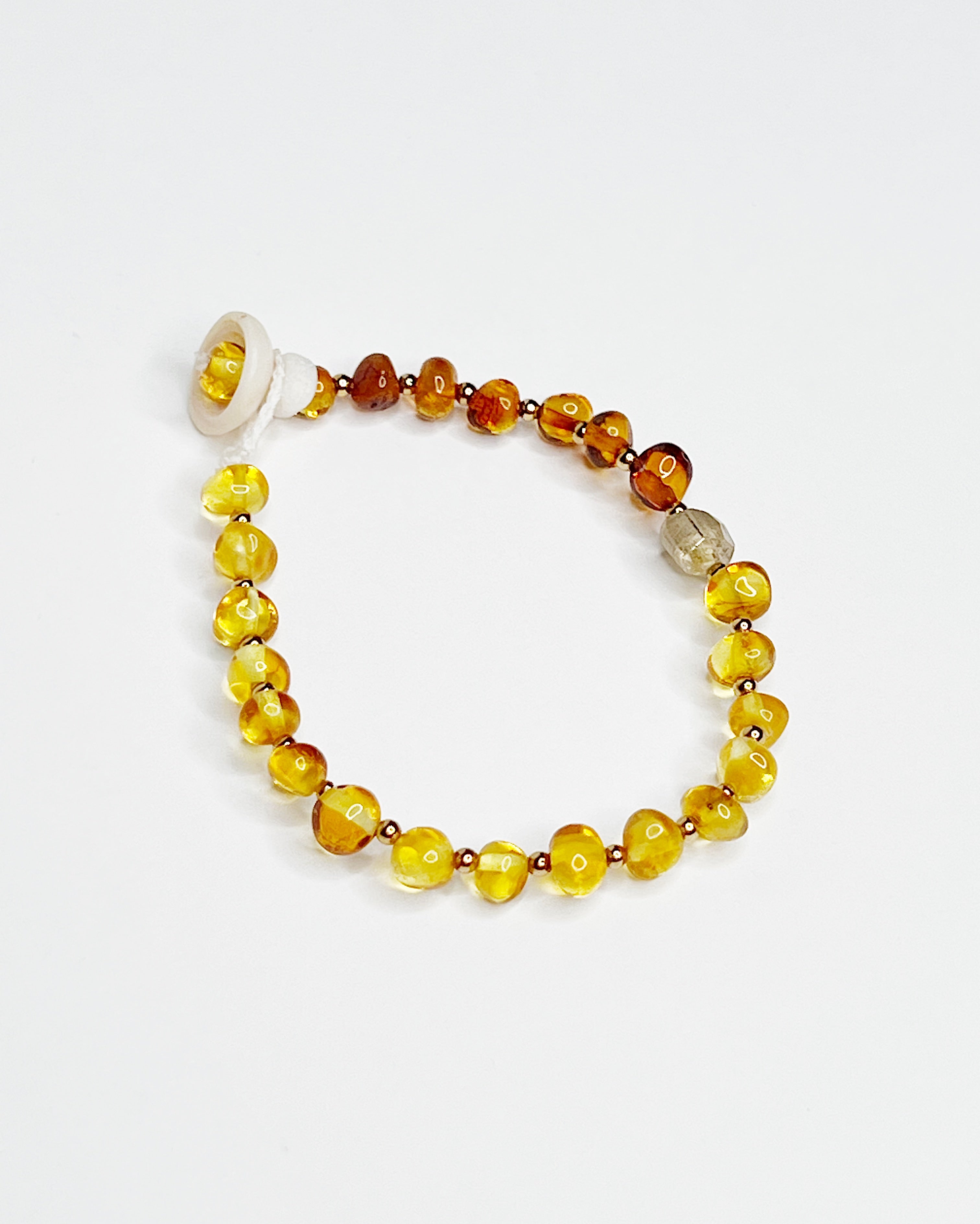 Handmade bracelet featuring polished Baltic amber beads in warm honey and cognac tones, accented with tiny gold spacers and finished with a tourmaline crystal and natural puka shell clasp, displayed on a white background