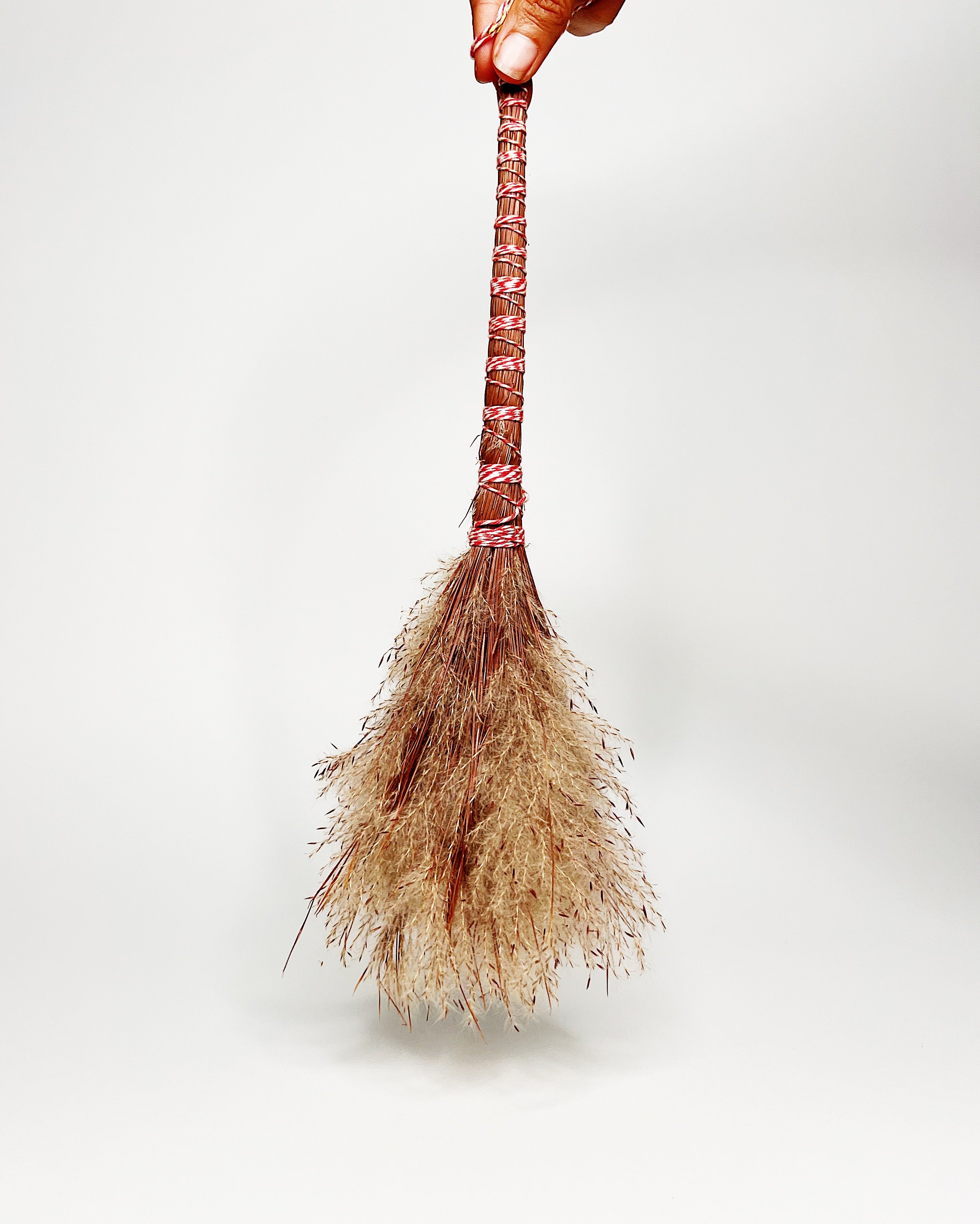 Traditional Hawaiian-style pūʻolo (broom) hand-tied with natural fibers and wrapped with red and white cord, featuring a tufted bundle of dried grass or sedge for sweeping. White background.