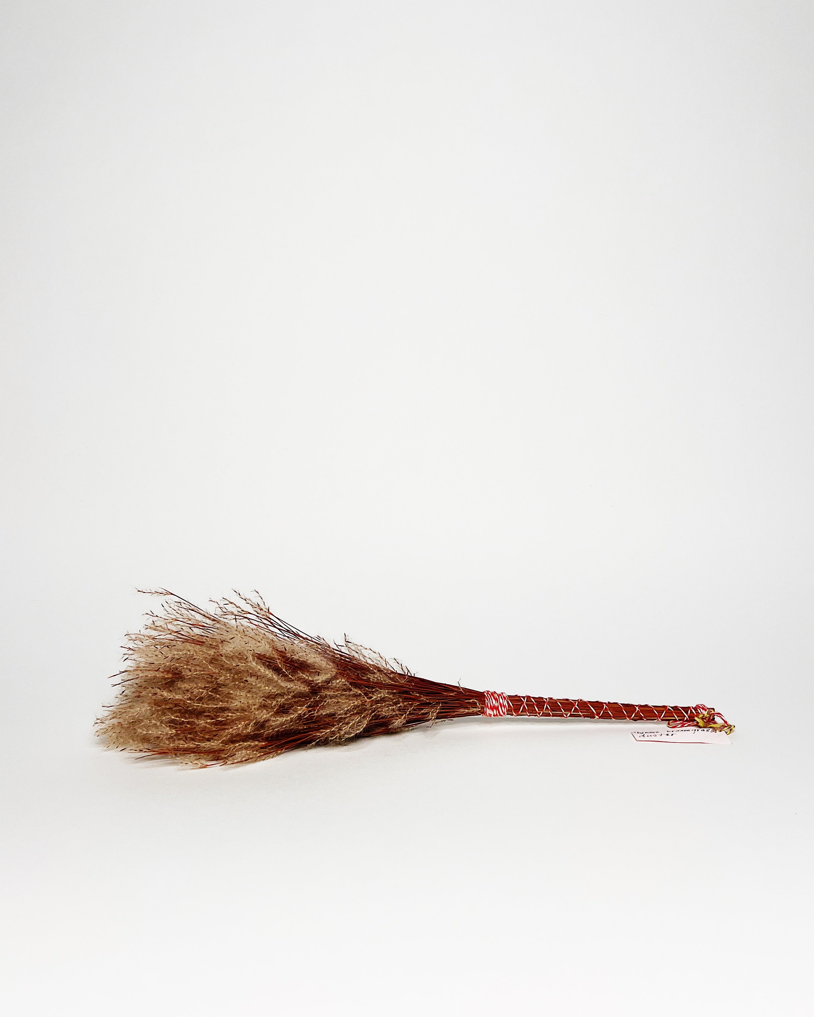 Traditional Hawaiian-style pūʻolo (broom) hand-tied with natural fibers and wrapped with red and white cord, featuring a tufted bundle of dried grass or sedge for sweeping. White background.