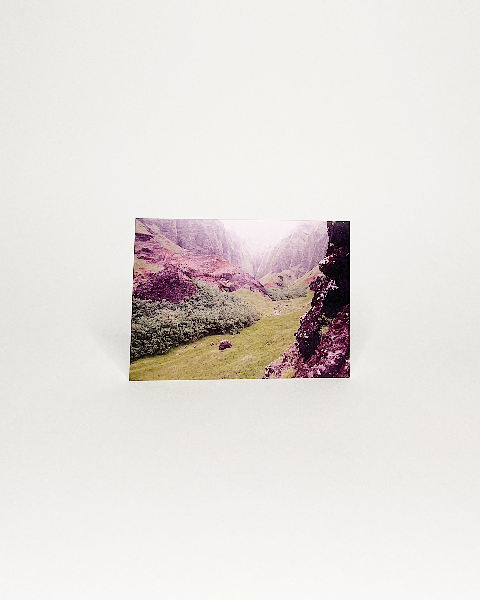 Photographic greeting card featuring a lush green valley framed by steep, rugged cliffs along Kauaʻi’s Nā Pali Coast, with soft mist settling at the distant ridgeline. White background