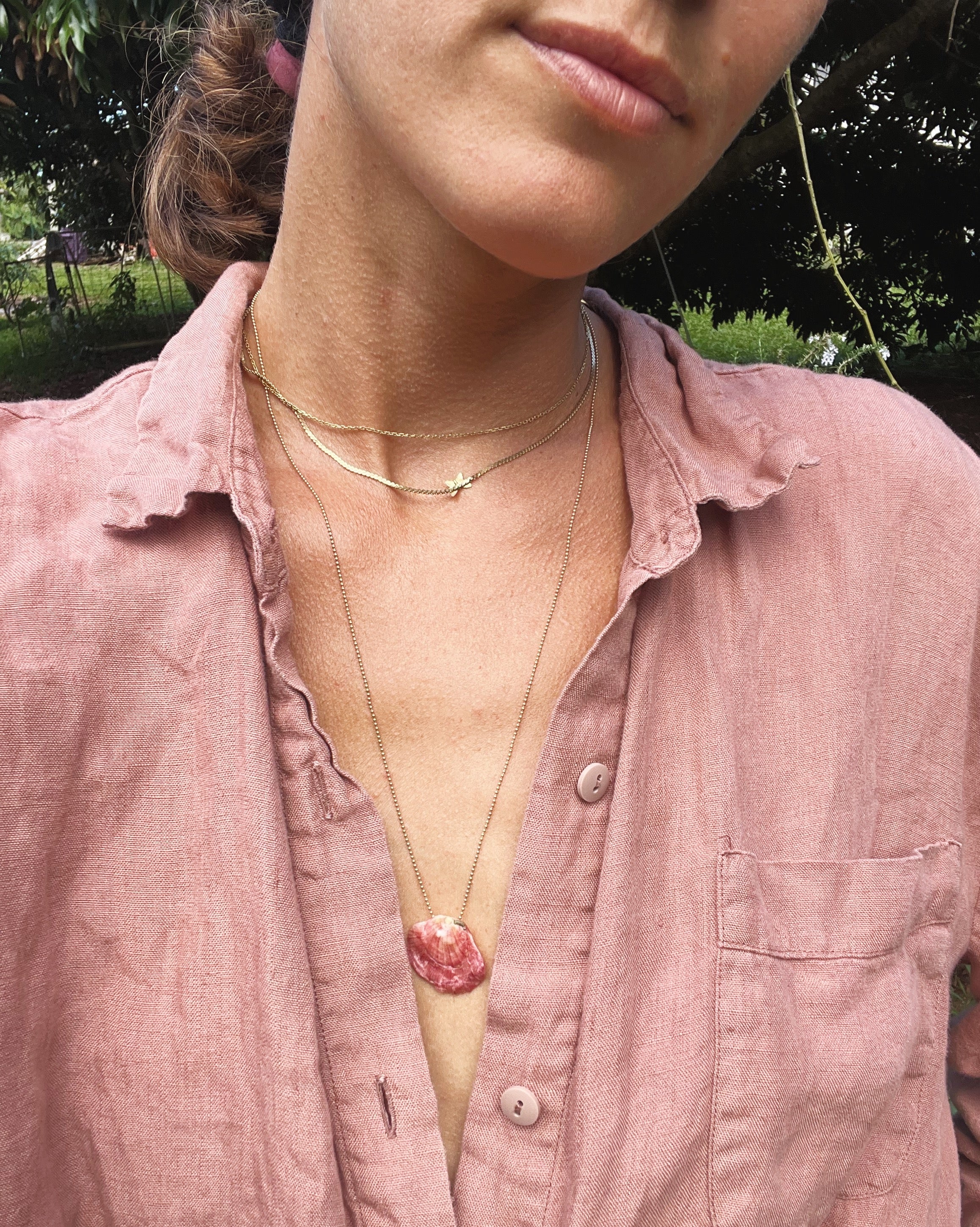 Woman wearing layered gold necklaces, featuring a pink shell pendant, styled with mauve button-down shirt