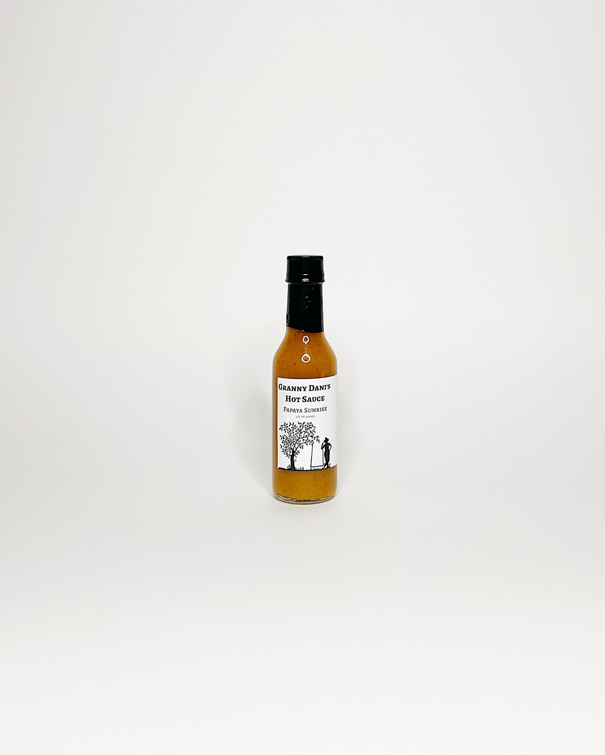 Bottle of Granny Dani’s Hot Sauce, Papaya Sunrise flavor, featuring a white label with a black silhouette of a tree and a figure, set against a white background.