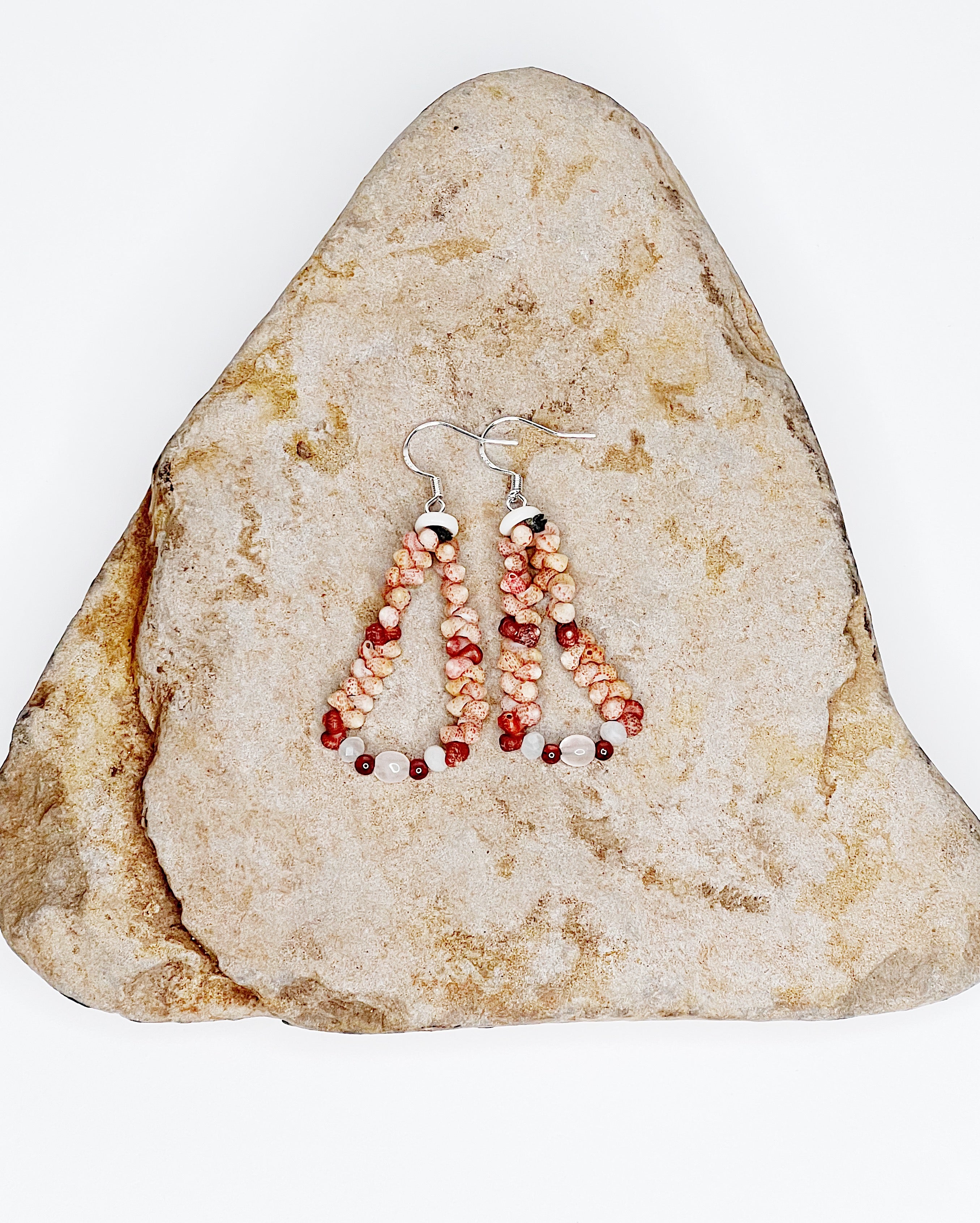 Handmade earrings featuring pink and red Kahelelani shells woven into a teardrop shape, accented with small moonstone beads at the base and finished with sterling silver hooks. Rock background