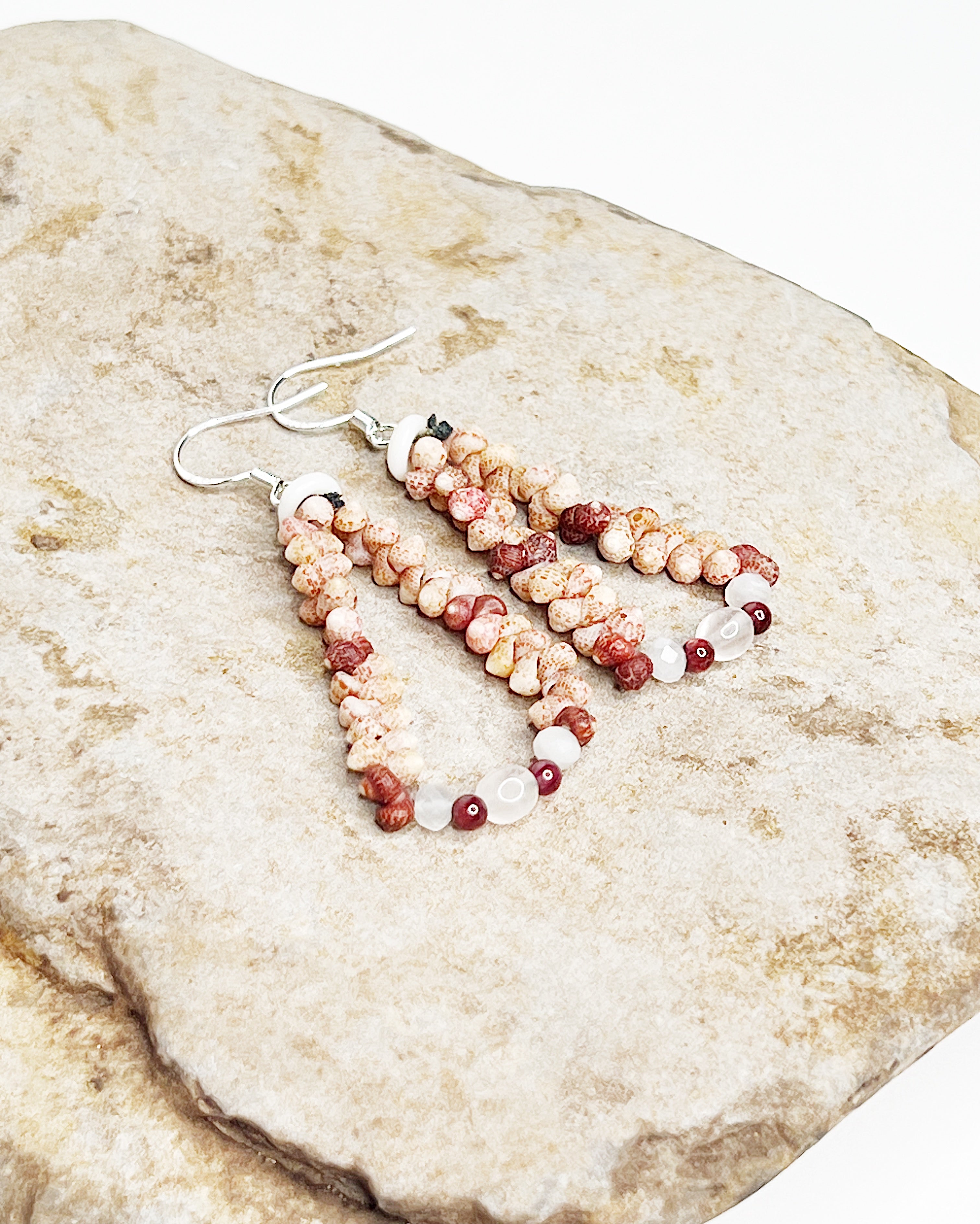 Handmade earrings featuring pink and red Kahelelani shells woven into a teardrop shape, accented with small moonstone beads at the base and finished with sterling silver hooks. Rock background