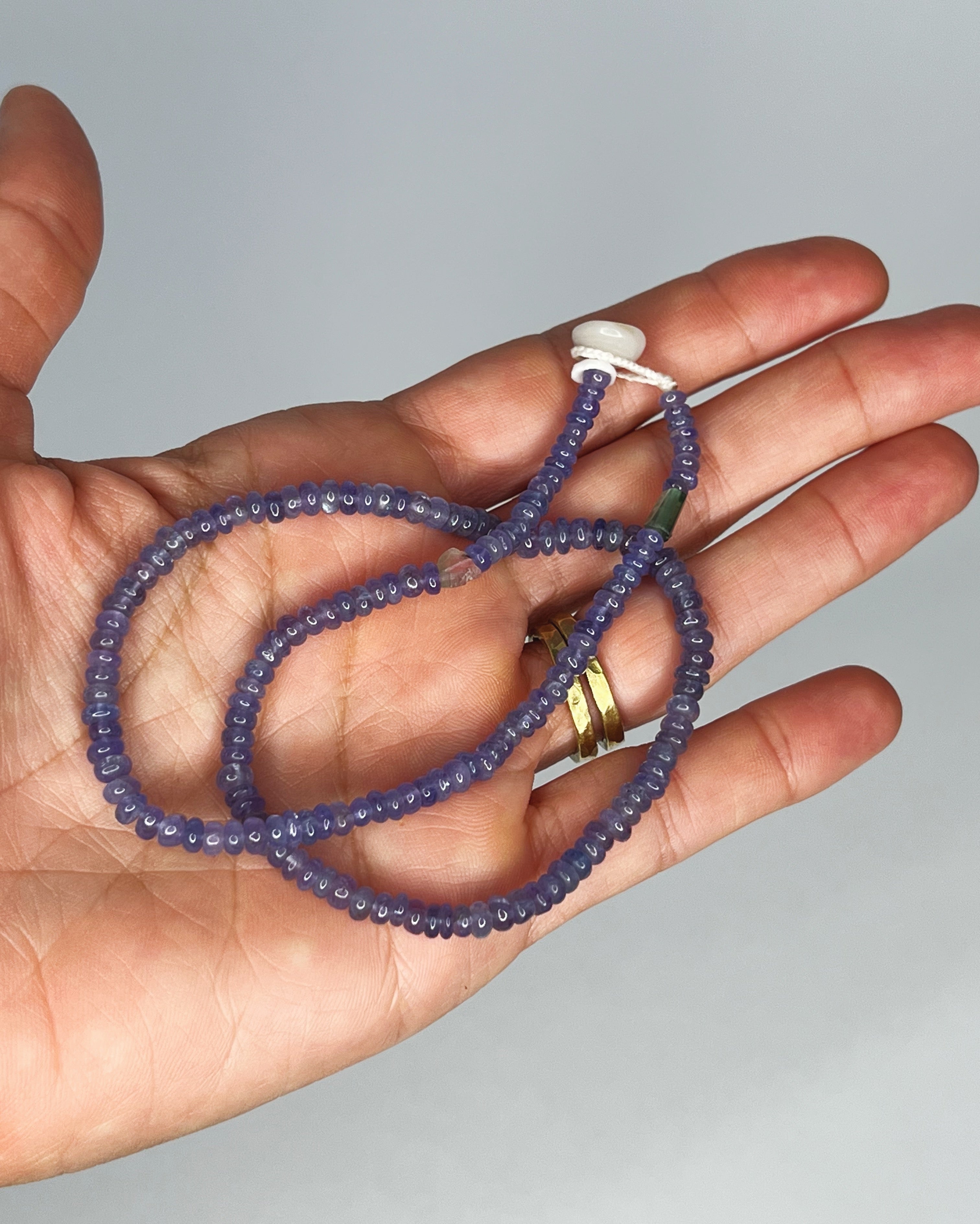 Hand holding Handmade tanzanite choker with tourmaline accents, finished with puka shell clasp. White background