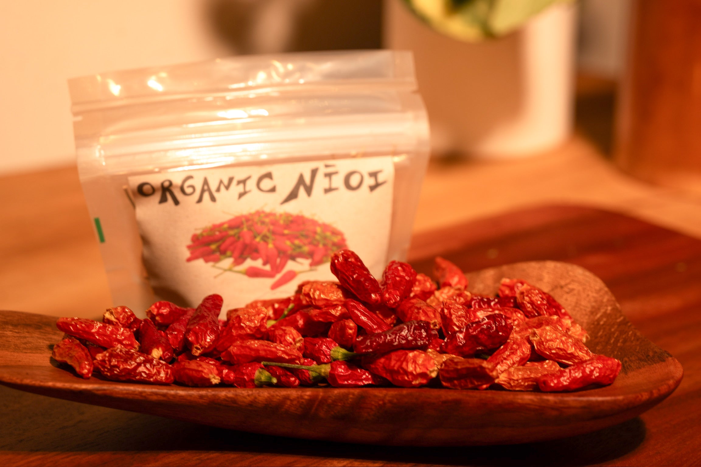 Close-up of dried organic Nīoi (Hawaiian chili peppers) in a wooden bowl, with a labeled resealable pouch in the background from ‘Ohi ‘Ohi Shop on Kaua‘i