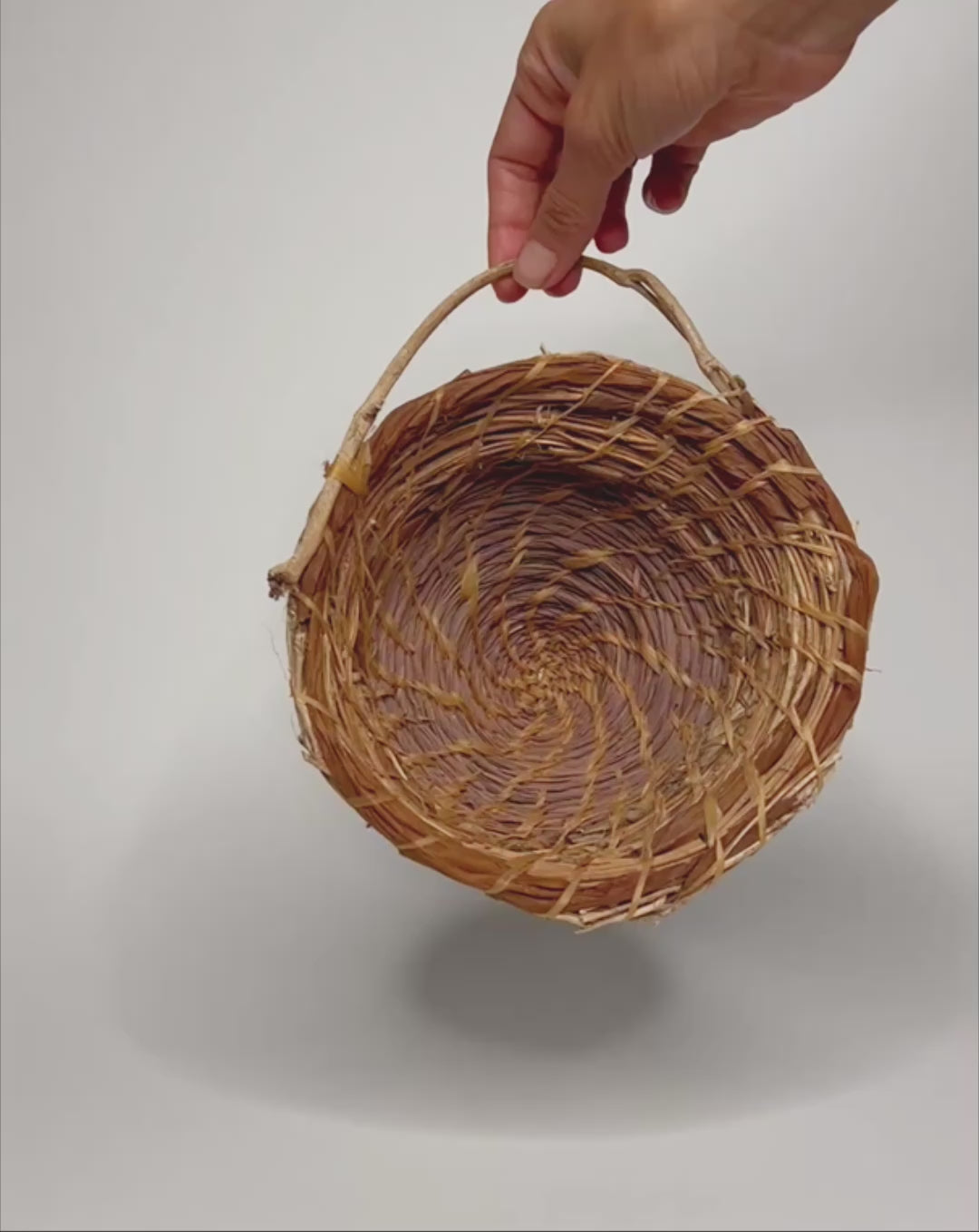 Movie of hand holding and turning a Handwoven basket crafted from lemongrass, lauaʻe fern, morning glory vine, and hau fiber, featuring a natural spiral weave pattern and a small organic handle, displayed against a clean white background