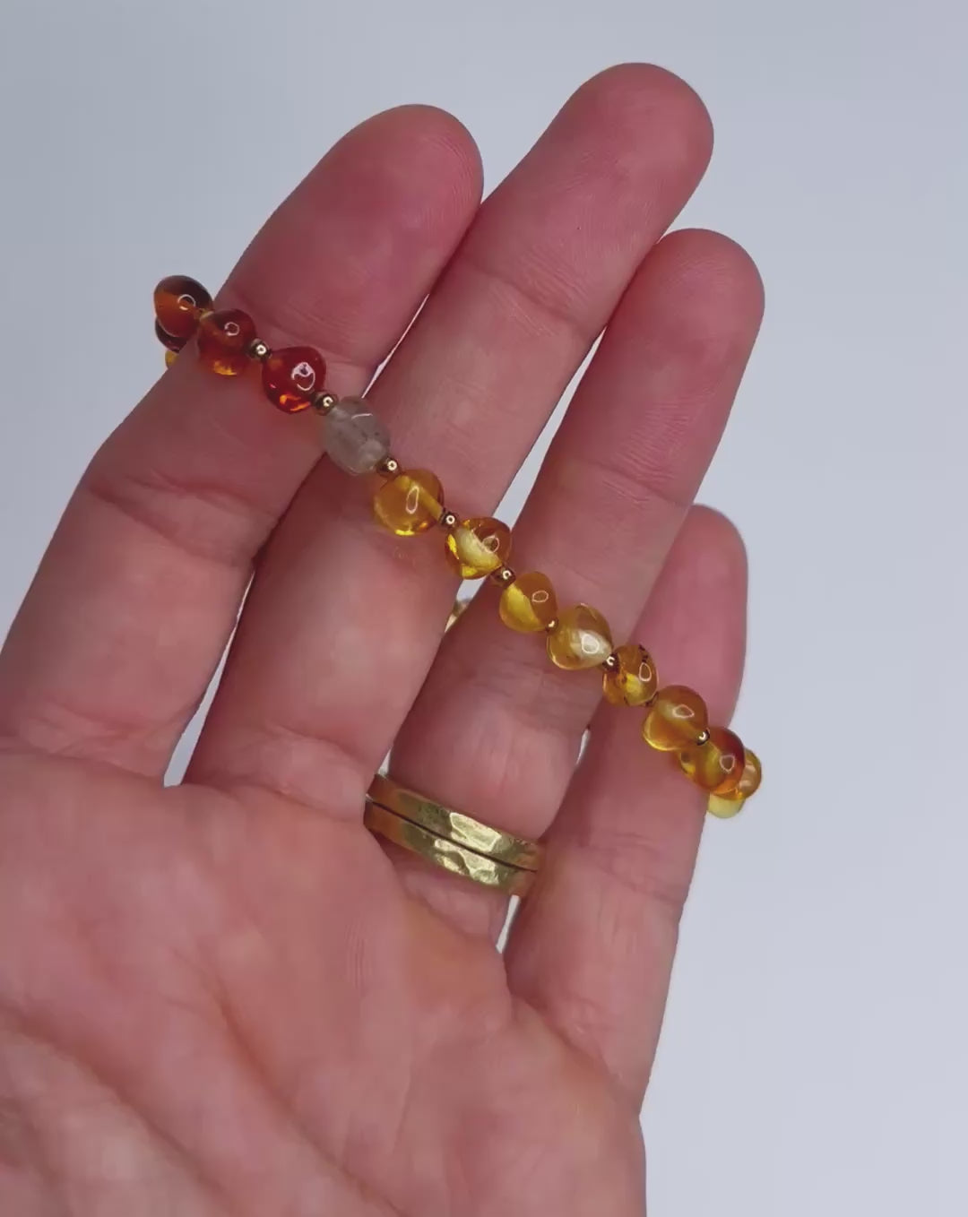 Video of hand showing Handmade bracelet featuring polished Baltic amber beads in warm honey and cognac tones, accented with tiny gold spacers and finished with a tourmaline crystal and natural puka shell clasp, displayed on a white background