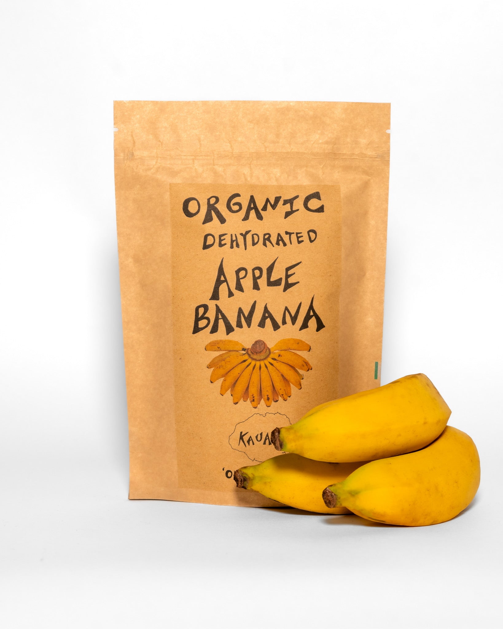 Organic dehydrated apple banana snack in kraft pouch with fresh Kaua‘i apple bananas, from ‘Ohi ‘Ohi Sho. Bananas laying aside the pouch. White background 