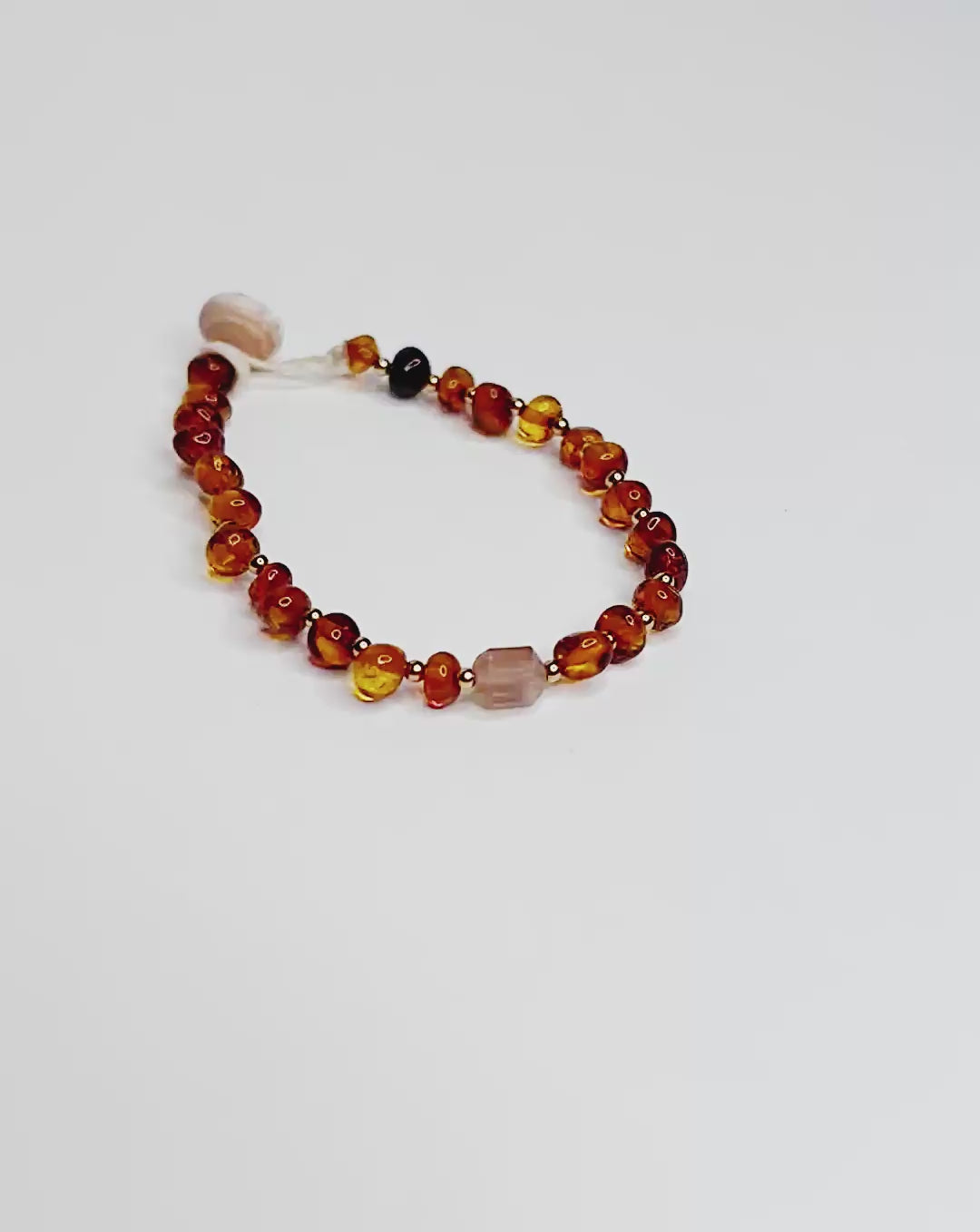 Video of Handmade bracelet featuring polished Baltic amber beads in cognac tones, accented with tiny gold spacers and finished with a tourmaline crystal and natural puka shell clasp, displayed on a white background