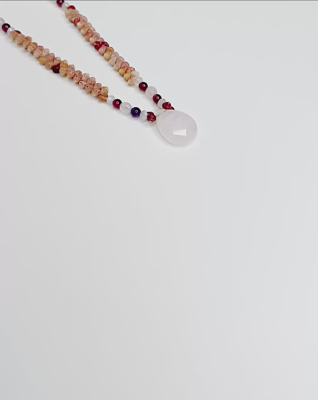 Movie of Handmade necklace featuring natural Kauaʻi Kahelelani shells intricately woven together, accented with garnet and moonstone beads and a faceted rose quartz teardrop pendant at the center, displayed against a white background